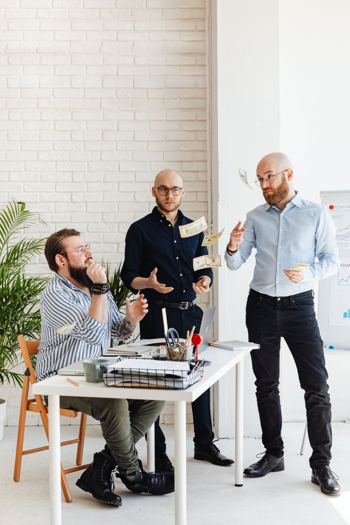 Three men discuss business plans at an office desk, tossing papers and exchanging ideas.