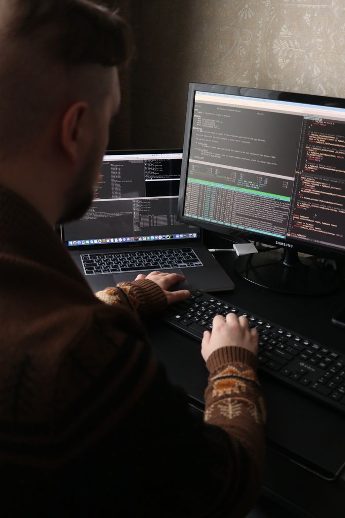 A programmer working on code with dual monitors and a laptop in a dimly lit room.